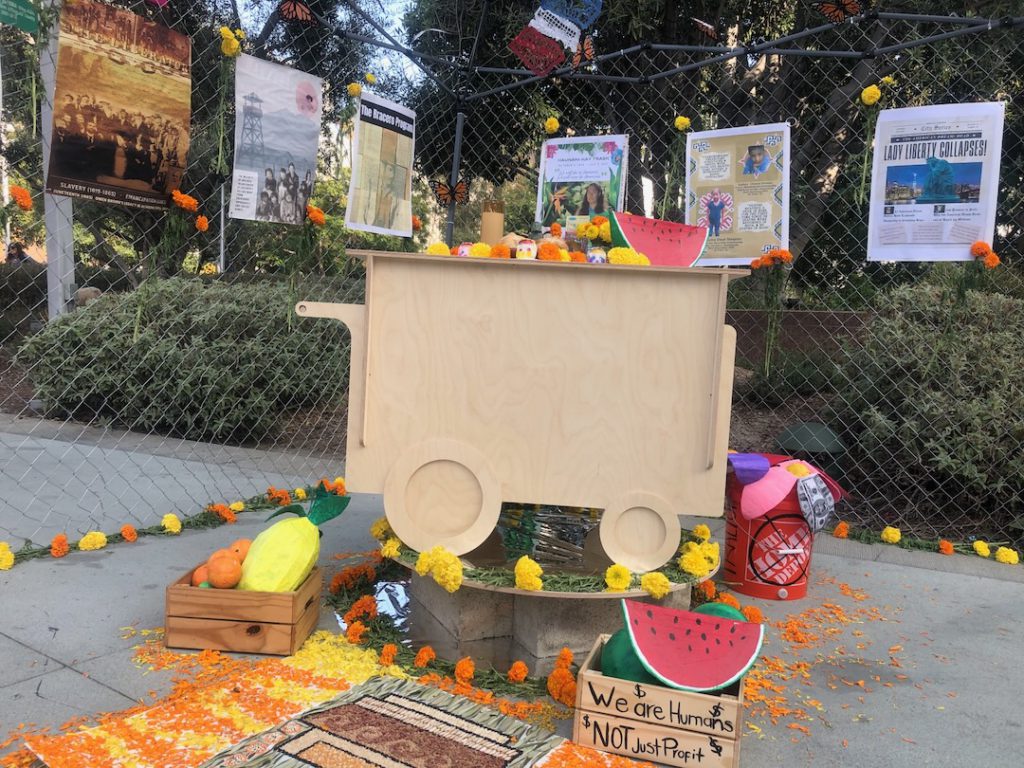 Dia de los Muertos altar "Death of the American Dream" by Danza Divina de Los Angeles includes sculpture of a fruit cart with sculpted fruits, including watermelon, a chain link fence with posters addressing immigration and indigenous issues and honoring those who have died and flowers in a Home Depot bucket (Photo: Liz Ohanesian)
