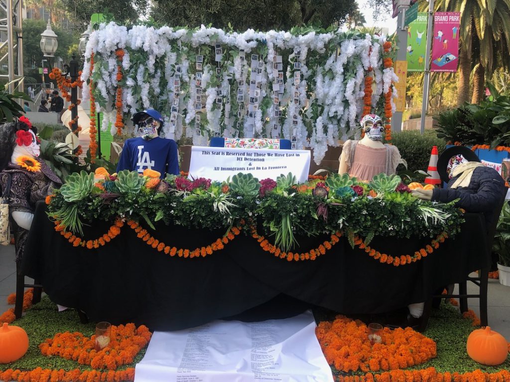 "Estamos Unidos" by LADWP- Society of Hispanic Professional Engineers. Altar at Gloria Molina Grand Park depicts skeletons at a family dining table with one empty seat with a sign that reads "This Seat Is Reserved for Those We Lost in ICE Detentions and all immigrants lost in la frontera" (Photo: Liz Ohanesian)