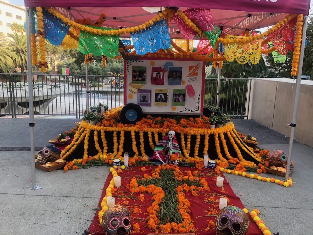 "Our Spirit Never Dies" by The Wall Las Memorias Staff and Volunteers at Gloria Molina Grand Park Altar features an ice cream cart with photos of those who have died in 2025. It is surrounded by flowers and a cross made of flowers lays on the ground in front of the altar. (Photo: Liz Ohanesian)