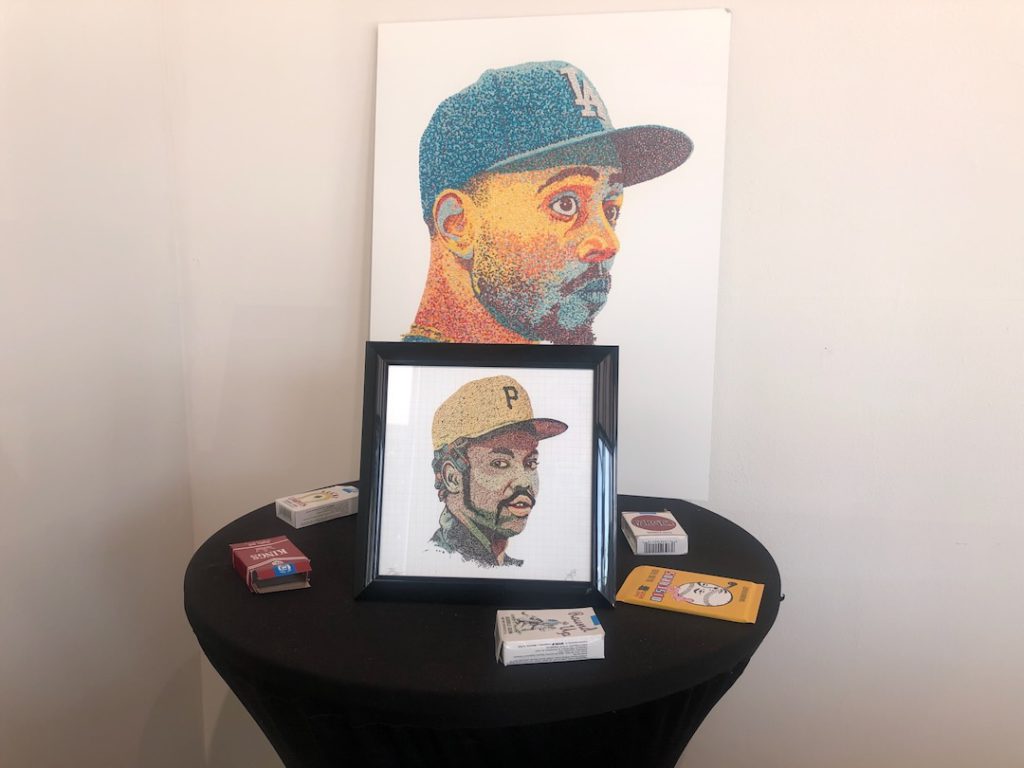 Print of Mookie Betts bubblegum portrait by Pat Riot behind a table with gum cigarettes on it. On the table is a print of a bubblegum portrait of Dock Ellis. (Photo: Liz Ohanesian)