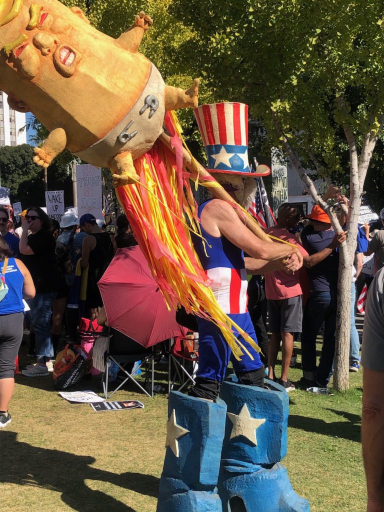A man dressed as Uncle Sam in massive platform shoes waves a sculpted sign of Trump as a baby at No Kings in downtown Los Angeles on October 18, 2025 (Photo: Liz Ohanesian)
