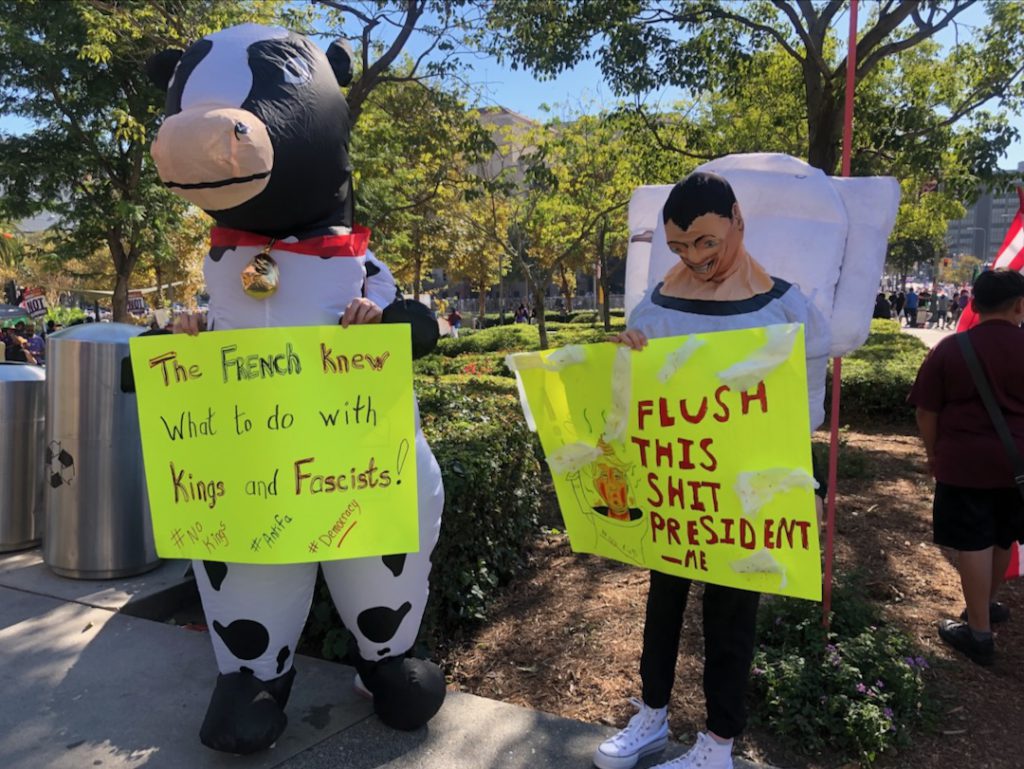 A protestor in an inflatable cow costume holds a sign that reads "The French knew what to do with kings and fascists." A protestor dressed as a toilet holds a sign that reads "Flush this shit president" (Photo: Liz Ohanesian)
