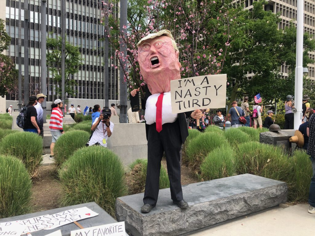 Protestor at No Kings in downtown Los Angeles dressed as Donald Trump with an oversized head mask holding a sign that reads, "I'm a nasty turd" (Photo: Liz Ohanesian)